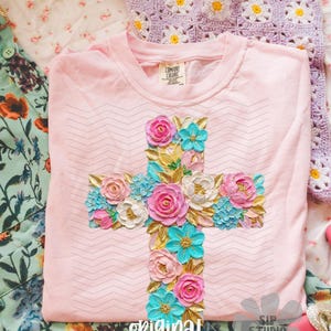May include: A light pink t-shirt with a floral cross design. The cross is adorned with pink, white, and turquoise flowers, and gold-colored leaves. The text "original designer" is at the bottom of the shirt.