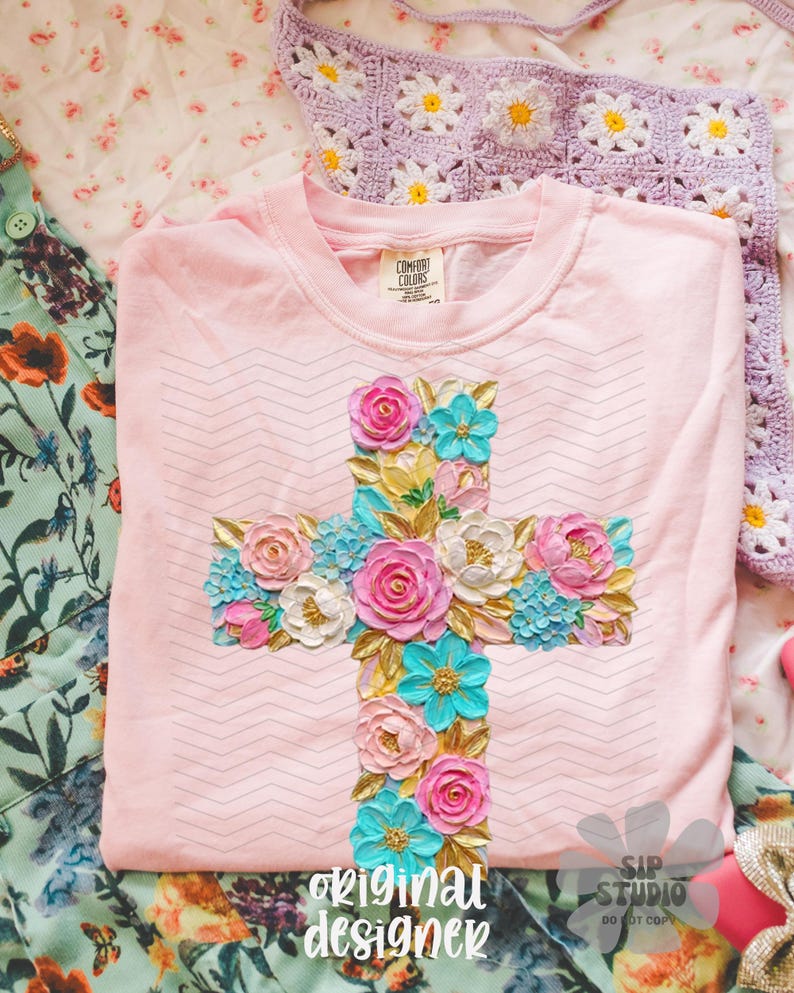 May include: A light pink t-shirt with a floral cross design. The cross is adorned with pink, white, and turquoise flowers, and gold-colored leaves. The text "original designer" is at the bottom of the shirt.