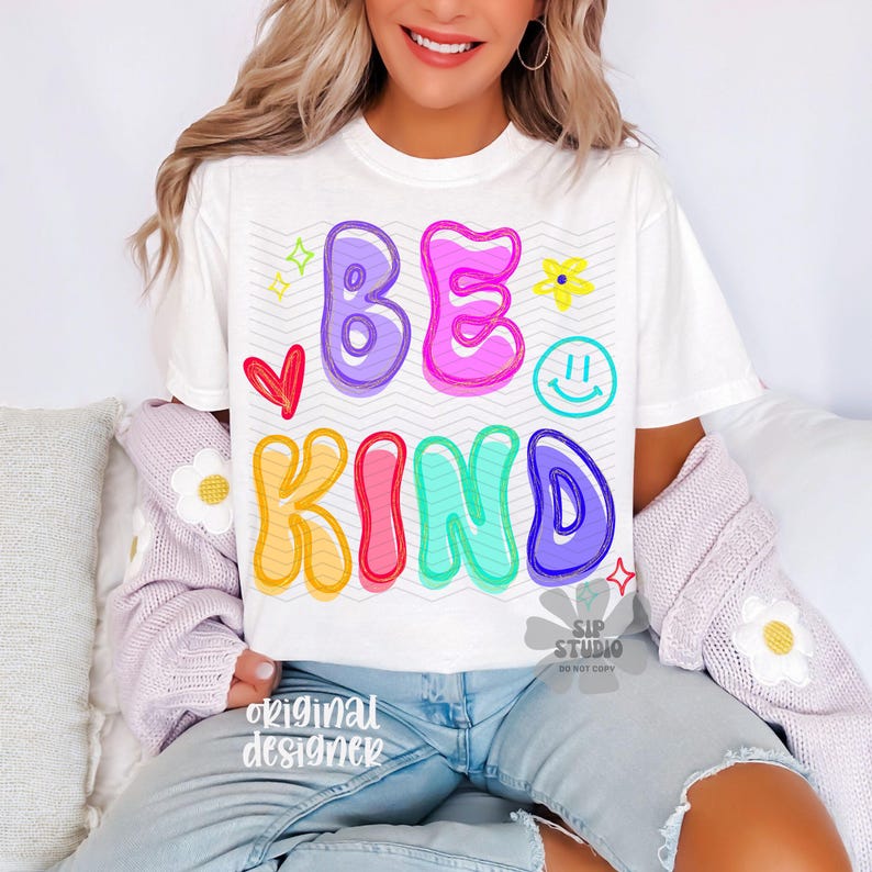 May include: White t-shirt with the words "BE KIND" in colorful bubble letters. The design includes a red heart, a yellow flower, a smiley face, and stars. The shirt is part of a casual wear collection.