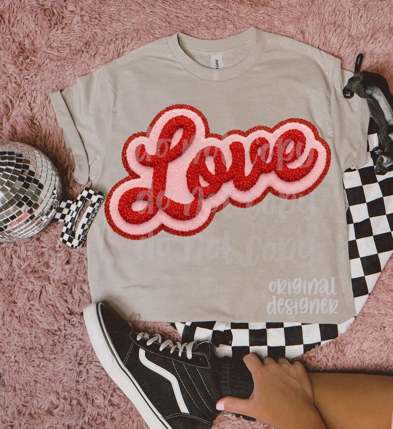 May include: A light tan t-shirt with the word "Love" in a red, textured font with a pink outline. The shirt is paired with black and white checkered pants and black sneakers.