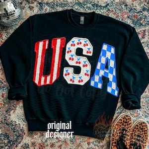 May include: Black sweatshirt with the word "USA" in red and white stripes, cherries, and blue and white checkered patterns. The words "original designer" are printed at the bottom.