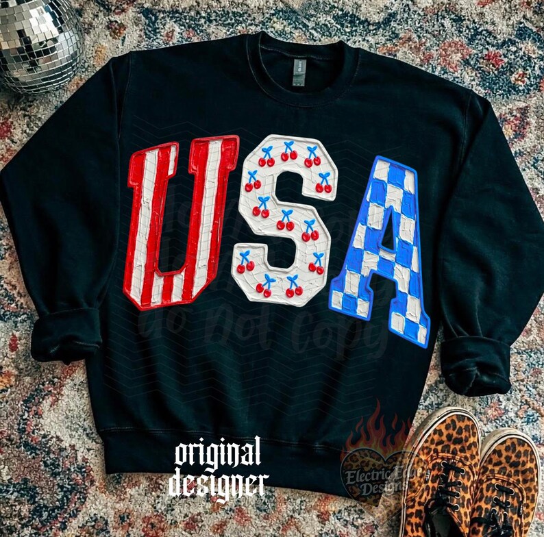 May include: Black sweatshirt with the word "USA" in red and white stripes, cherries, and blue and white checkered patterns. The words "original designer" are printed at the bottom.
