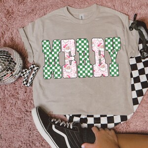 May include: A beige t-shirt with the word "MERRY" in a checkerboard design. The letters "M" and "Y" are green and white, while the "E" and "R" are pink with Santa Claus faces. The shirt is folded.