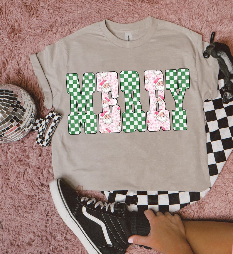 May include: A beige t-shirt with the word "MERRY" in a checkerboard design. The letters "M" and "Y" are green and white, while the "E" and "R" are pink with Santa Claus faces. The shirt is folded.
