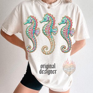 May include: Cream-colored t-shirt featuring three colorful seahorse illustrations. The seahorses have teal, pink, and gold accents. The shirt also has the text "original designer" in a gothic font and a logo.