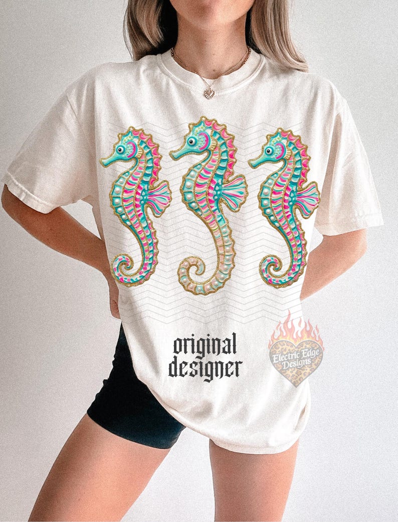 May include: Cream-colored t-shirt featuring three colorful seahorse illustrations. The seahorses have teal, pink, and gold accents. The shirt also has the text "original designer" in a gothic font and a logo.