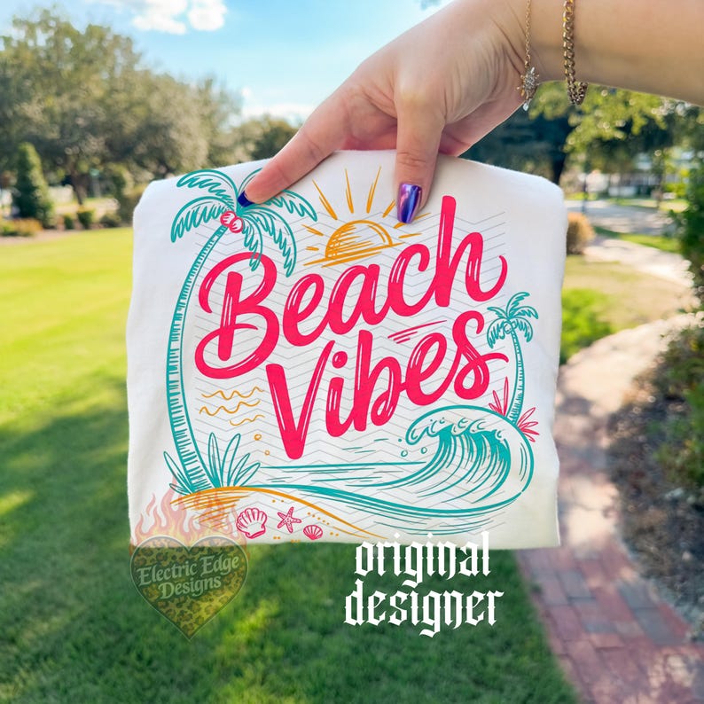 May include: White t-shirt with a colorful "Beach Vibes" graphic. The design includes a sun, palm trees, waves, and the text "Beach Vibes" in pink and teal. The shirt is held up by a hand.