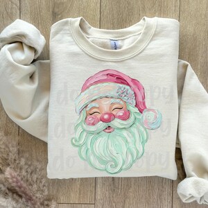May include: Cream-colored sweatshirt featuring a Santa Claus graphic. The Santa has a pink hat, a mint green beard, and rosy cheeks. The sleeves are rolled up, and the sweatshirt is laid on a wooden surface.