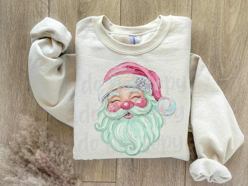 May include: Cream-colored sweatshirt featuring a Santa Claus graphic. The Santa has a pink hat, a mint green beard, and rosy cheeks. The sleeves are rolled up, and the sweatshirt is laid on a wooden surface.