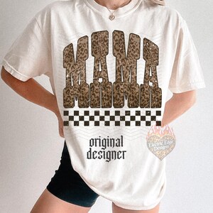 May include: Cream-colored t-shirt with the word "MAMA" in a leopard print design. Below the word "MAMA" is a black and white checkered pattern. The words "original designer" are printed below the checkered pattern.