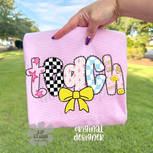 May include: A light pink t-shirt with the word "teach" in a colorful, playful design. The letters feature stars, checkered patterns, lightning bolts, and a yellow bow. The shirt is held up by a hand.