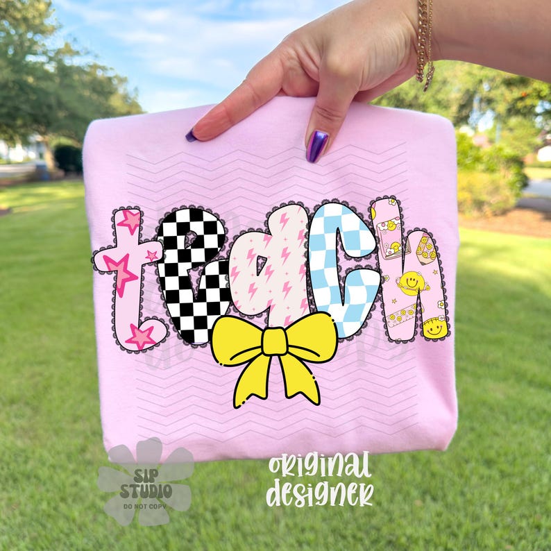 May include: A light pink t-shirt with the word "teach" in a colorful, playful design. The letters feature stars, checkered patterns, lightning bolts, and a yellow bow. The shirt is held up by a hand.