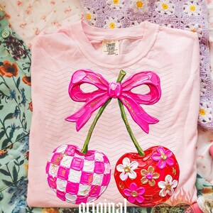May include: A light pink t-shirt featuring a graphic of two cherries connected by a bright pink bow. One cherry has a pink and white checkered pattern, while the other is red with white and gold floral accents. The design has a textured paint effect.
