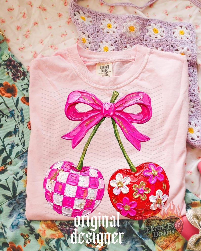 May include: A light pink t-shirt featuring a graphic of two cherries connected by a bright pink bow. One cherry has a pink and white checkered pattern, while the other is red with white and gold floral accents. The design has a textured paint effect.