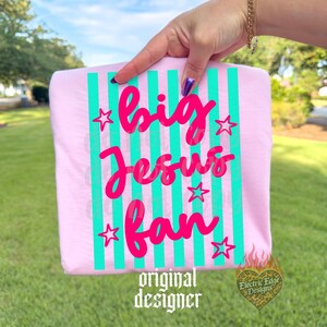 May include: A light pink t-shirt with vertical turquoise stripes and the words "big Jesus fan" in bright pink script. The shirt also features pink star accents and the words "original designer".