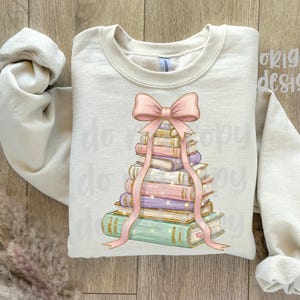 May include: Cream-colored sweatshirt featuring a stack of books with a pink bow and ribbon design. The books are in various colors, including green, purple, and gold. The words "original designer" are visible on the top right.