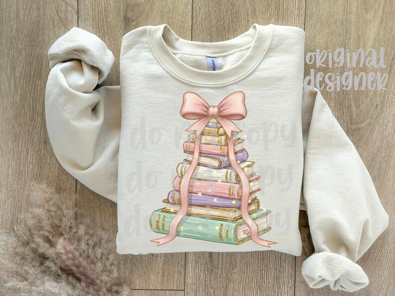 May include: Cream-colored sweatshirt featuring a stack of books with a pink bow and ribbon design. The books are in various colors, including green, purple, and gold. The words "original designer" are visible on the top right.