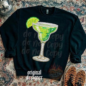 May include: Black sweatshirt featuring a sequined margarita glass with a lime wedge. The words "do not copy" are printed repeatedly on the shirt. The words "original designer" and a logo are also visible.