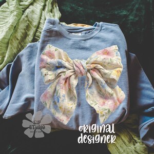 May include: A light blue sweatshirt with a large floral bow. The bow features a pastel floral pattern with pink, yellow, and blue flowers. The words "original designer" are printed in white script on the bottom right.