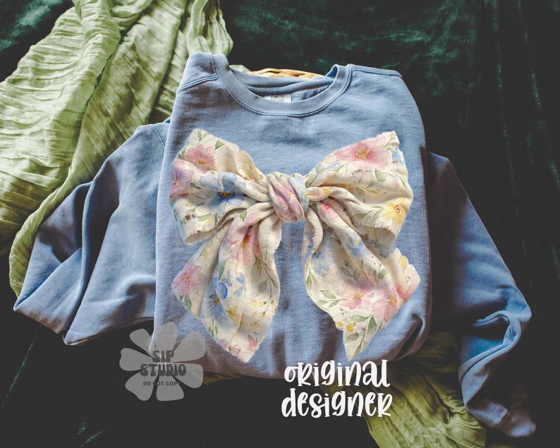 May include: A light blue sweatshirt with a large floral bow. The bow features a pastel floral pattern with pink, yellow, and blue flowers. The words "original designer" are printed in white script on the bottom right.