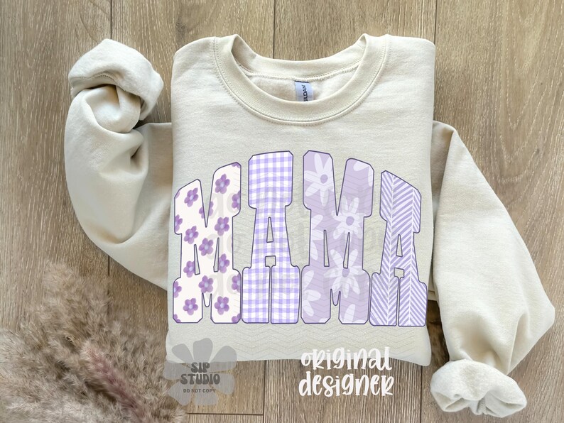 May include: A cream-colored sweatshirt with the word "MAMA" in large, purple letters. The letters have floral, gingham, and striped patterns. The words "SIP STUDIO" and "original designer" are also visible.
