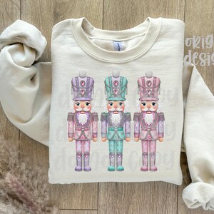 May include: A cream-coloured sweatshirt displaying three colourful nutcracker soldier designs. Each nutcracker has a detailed hat and uniform in shades of pink, purple, and teal. The words "original designer" are printed on the upper right.