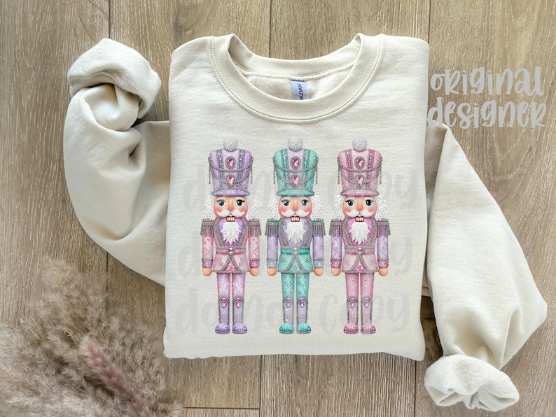May include: A cream-coloured sweatshirt displaying three colourful nutcracker soldier designs. Each nutcracker has a detailed hat and uniform in shades of pink, purple, and teal. The words "original designer" are printed on the upper right.
