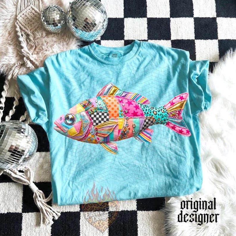 May include: A light blue t-shirt featuring a colorful, patchwork-style fish design. The fish incorporates various patterns, including stars, leopard print, and checkered squares, in shades of pink, orange, yellow, and blue. The words "original designer" are printed on the shirt.