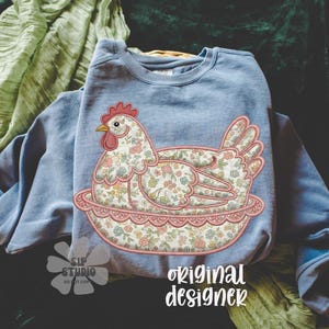 May include: A light blue sweatshirt featuring an embroidered hen design. The hen is adorned with a floral pattern in shades of pink, blue, and yellow. The design includes the text "original designer".