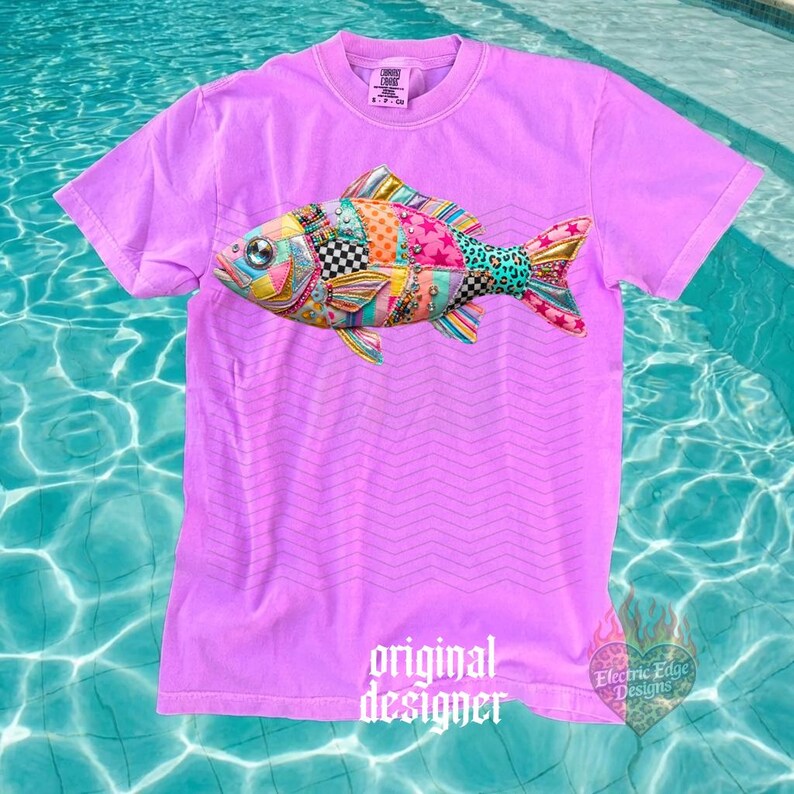 May include: A lilac-colored t-shirt featuring a colorful patchwork fish design. The fish has a variety of patterns and colors, including pink, blue, yellow, and black. The shirt also has the words "original designer" printed on it.