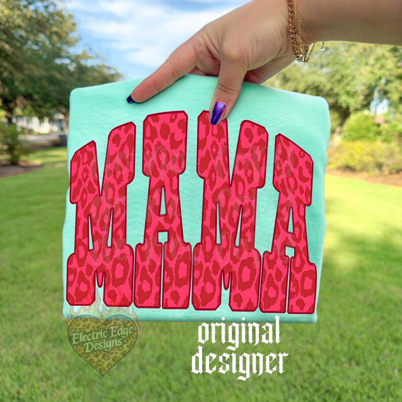 May include: A light mint green t-shirt featuring the word MAMA in large, bold, block letters. The letters have a vibrant pink and red leopard print pattern. A small heart-shaped logo with the text Electric Edge Designs is visible.