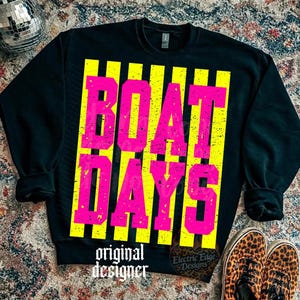 Może przedstawiać: Czarna bluza z napisem "BOAT DAYS" w jaskrawor&oacute;żowych literach na ż&oacute;łtych pionowych paskach. Słowa "original designer" są wydrukowane na dole na biało. Buty w panterkę widoczne w prawym dolnym rogu.