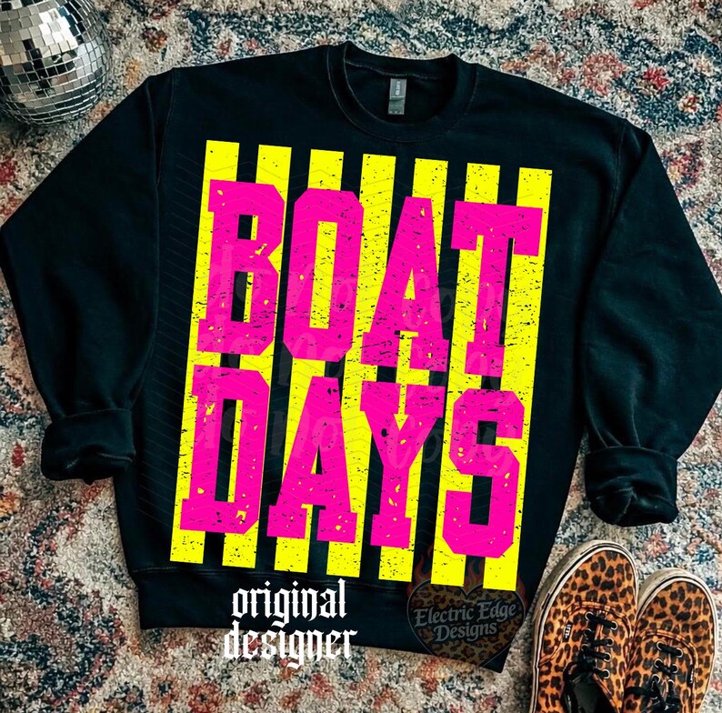 Może przedstawiać: Czarna bluza z napisem "BOAT DAYS" w jaskrawor&oacute;żowych literach na ż&oacute;łtych pionowych paskach. Słowa "original designer" są wydrukowane na dole na biało. Buty w panterkę widoczne w prawym dolnym rogu.
