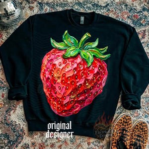 May include: Black sweatshirt featuring a large, detailed strawberry graphic. The strawberry is rendered in shades of red and pink with gold accents and green leaves. The text "original designer" is printed on the bottom.