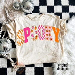 May include: A cream-colored t-shirt featuring the word SPOOKY in large, colorful letters. The letters display various patterns including orange and white checkers, solid hot pink, white with orange ghosts and flowers, pink and white checkers, and orange.