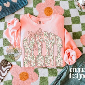 May include: A coral-colored sweatshirt with the word "MAMA" in floral print letters, accented with a bow. The design is set against a green and white checkered background with pink flower-shaped rugs. A phone case and jeans are also visible.