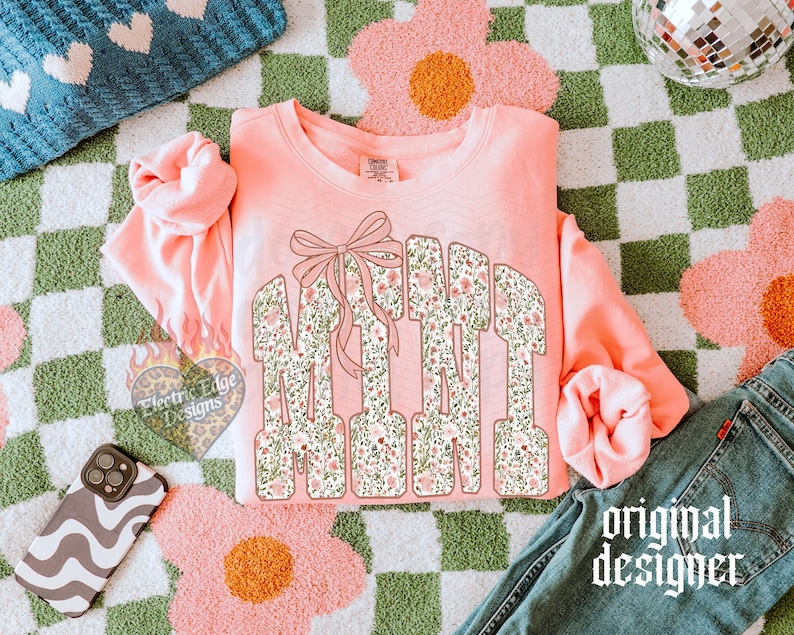 May include: A coral-colored sweatshirt with the word "MAMA" in floral print letters, accented with a bow. The design is set against a green and white checkered background with pink flower-shaped rugs. A phone case and jeans are also visible.