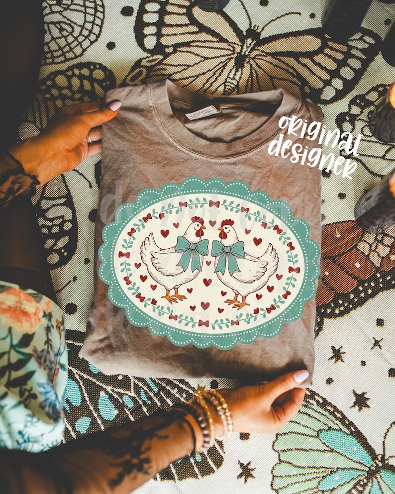 May include: A brown t-shirt featuring a whimsical design of two chickens with teal bows, surrounded by hearts and bows, within a teal oval frame. The words "original designer" are visible on the shirt.