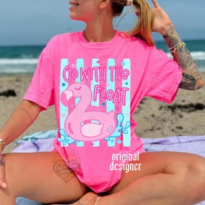 May include: A bright pink t-shirt with the text "Go With The Float" and a flamingo floatie design. The design features a pink flamingo, blue water splashes, and blue vertical stripes. The words "original designer" are also printed on the shirt.