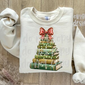 May include: Cream-colored sweatshirt featuring a Christmas tree design made of stacked books. The books are green and gold, adorned with string lights and topped with a red bow with white polka dots. The sleeves are rolled up.