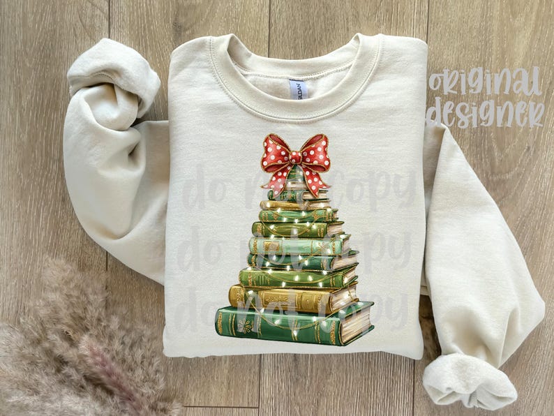 May include: Cream-colored sweatshirt featuring a Christmas tree design made of stacked books. The books are green and gold, adorned with string lights and topped with a red bow with white polka dots. The sleeves are rolled up.