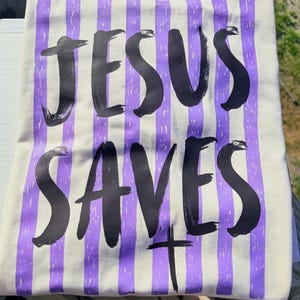 May include: Cream-colored t-shirt with vertical purple stripes. The words "JESUS SAVES" are printed in large, black, brush-style lettering. A black cross is below the word "SAVES".