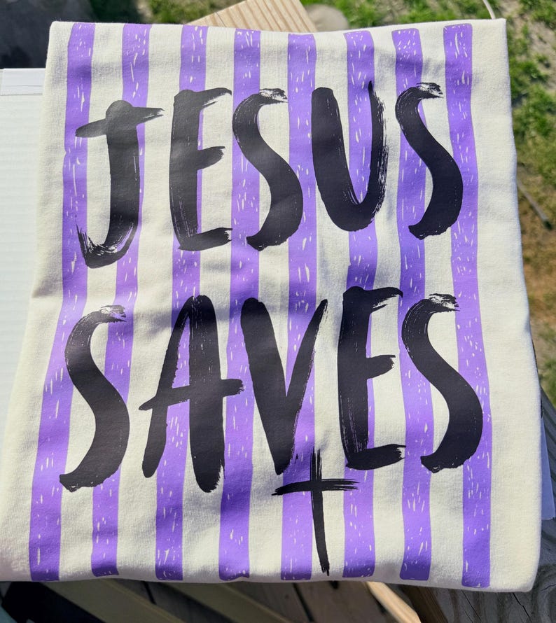 May include: Cream-colored t-shirt with vertical purple stripes. The words "JESUS SAVES" are printed in large, black, brush-style lettering. A black cross is below the word "SAVES".