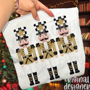 May include: White cushion cover with three nutcracker designs. Each nutcracker has a gold and black uniform, a black and white chequered hat with a gold star, and a handlebar moustache. The words "original designer" are in the bottom right corner.