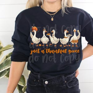 May include: Navy blue sweatshirt featuring a graphic of five cartoon geese, each with a different Thanksgiving-themed accessory. The text "just a thankful goose" is in orange script. The sweatshirt is a long-sleeved crew neck.