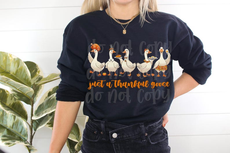 May include: Navy blue sweatshirt featuring a graphic of five cartoon geese, each with a different Thanksgiving-themed accessory. The text "just a thankful goose" is in orange script. The sweatshirt is a long-sleeved crew neck.
