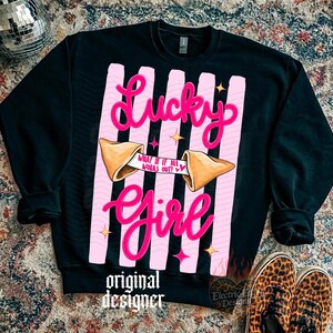 May include: Black sweatshirt featuring a pink and white design. The design includes the words "Lucky Girl" in a cursive font, a fortune cookie, and the text "What if it all works out?" The sweatshirt also has the words "original designer".