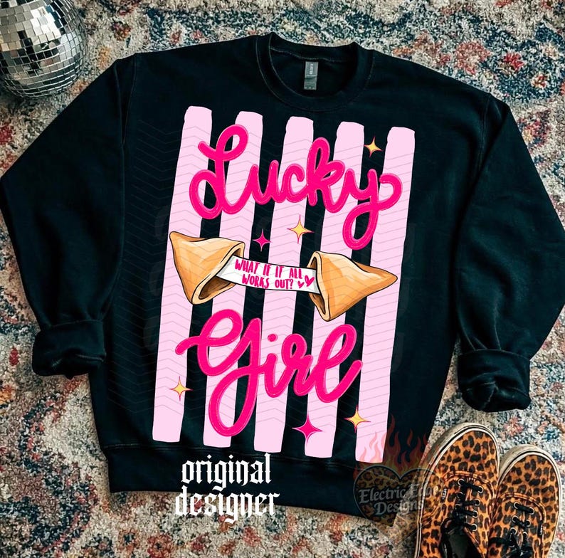 May include: Black sweatshirt featuring a pink and white design. The design includes the words "Lucky Girl" in a cursive font, a fortune cookie, and the text "What if it all works out?" The sweatshirt also has the words "original designer".