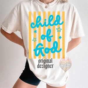 May include: Cream-colored t-shirt with the text "child of God" in turquoise, set against yellow and white vertical stripes. The shirt also features the words "original designer" in black, and a logo.