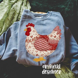 May include: A pale blue sweatshirt with a chicken appliqu&eacute;. The chicken design uses red and white floral and gingham patterns, with a red comb and yellow feet. The text "original designer" is at the bottom of the image.